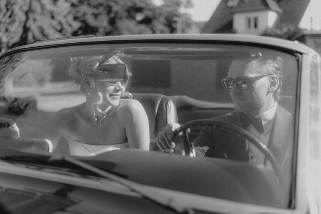 Brautpaar im Oldtimer mit Sonnenbrillen, stilvolle Hochzeitsfotografie in Schwarzweiß